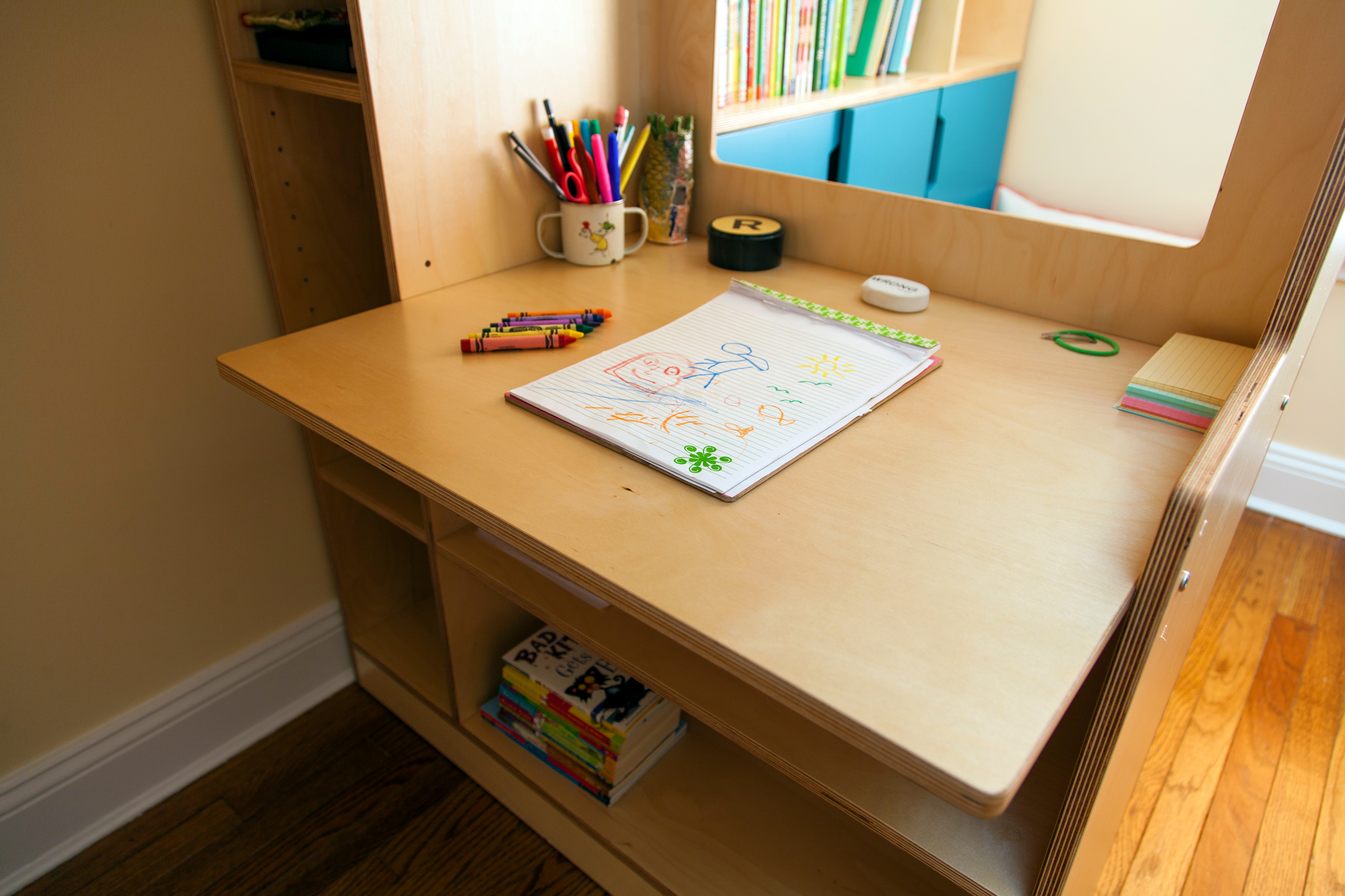 A small wooden study desk with colored pencils and a drawing book, positioned near shelves with books in a bright room.