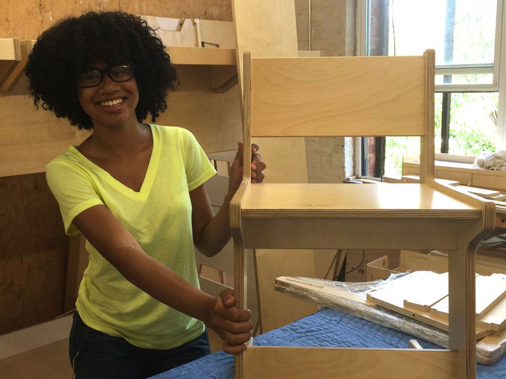 Woman in a yellow shirt proudly holding a wooden chair she built, standing in a workshop with a smile on her face.