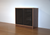 Minimalist wooden cabinet with decorative slatted doors on a polished hardwood floor against a white wall.