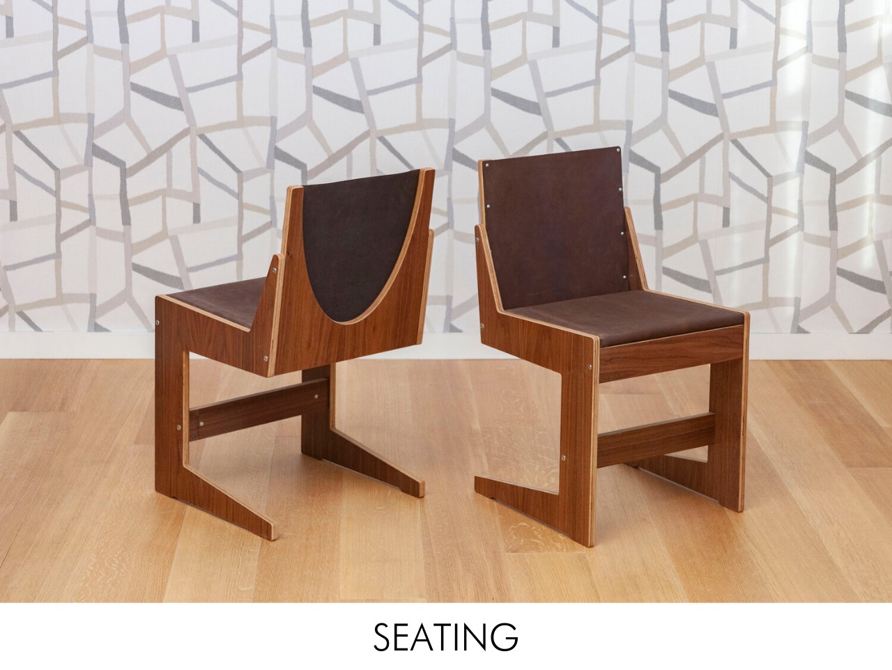 Two contemporary wooden chairs with leather backrests against a geometric patterned wallpaper.