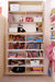 A tall bookshelf filled with dolls, books, and toys, alongside white wicker baskets for storage, with backpacks hanging nearby.