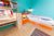 A vibrant kids' room with a turquoise wall, single bed with orange drawers, a desk, shelves, and a colorful striped rug.