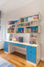 A modern study area with a large desk and bright blue drawers, featuring shelves with books, toys, and decorative items.