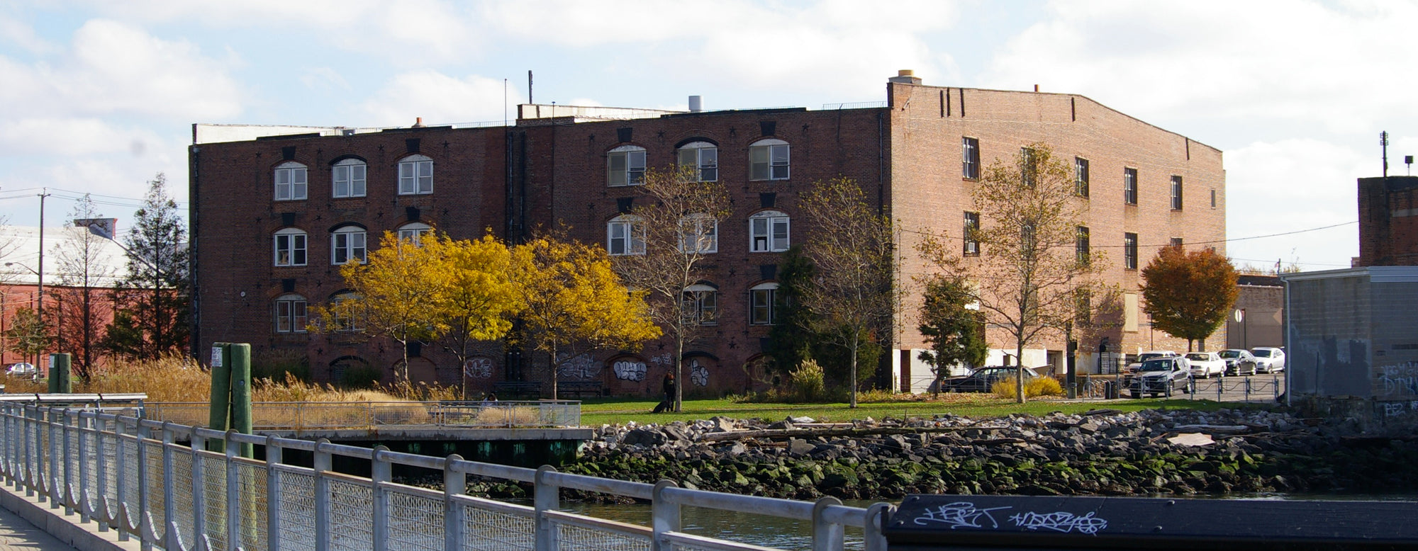 Old industrial brick building near a waterway with a railing and autumn trees.