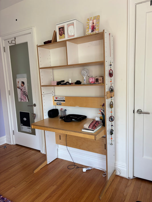 A light wood desk with an attached open shelving unit, shown in a home office setting with wood floors, white walls, and two doors.