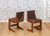 Two contemporary wooden chairs with black leather backrests on a hardwood floor against a geometric wallpaper.