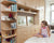 Child in polka dot top sits on bed, facing bookshelf and cabinets in a sunny room.