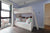 A kids' room with a bunk bed, plush toys, colorful bedding, light blue walls, a large window, and a door leading to another room.