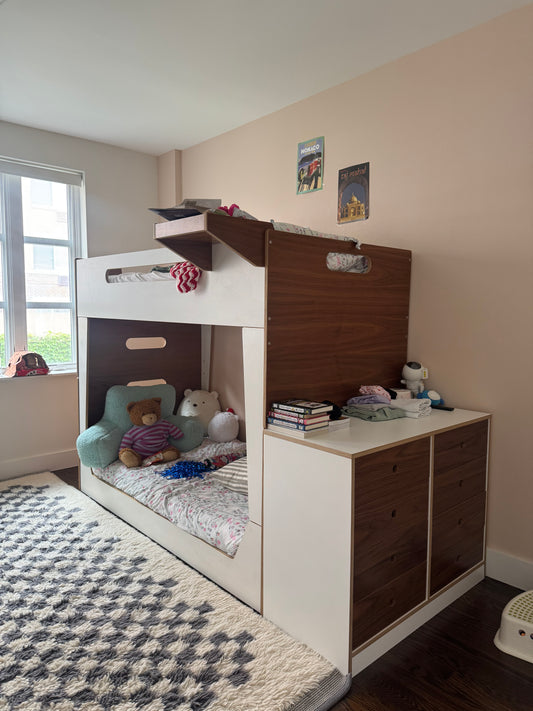 A bunk bed with a white frame and dark brown wood-grain surfaces, featuring a built-in desk, open storage shelf with toys, and a small bed underneath, shown in a bedroom setting.