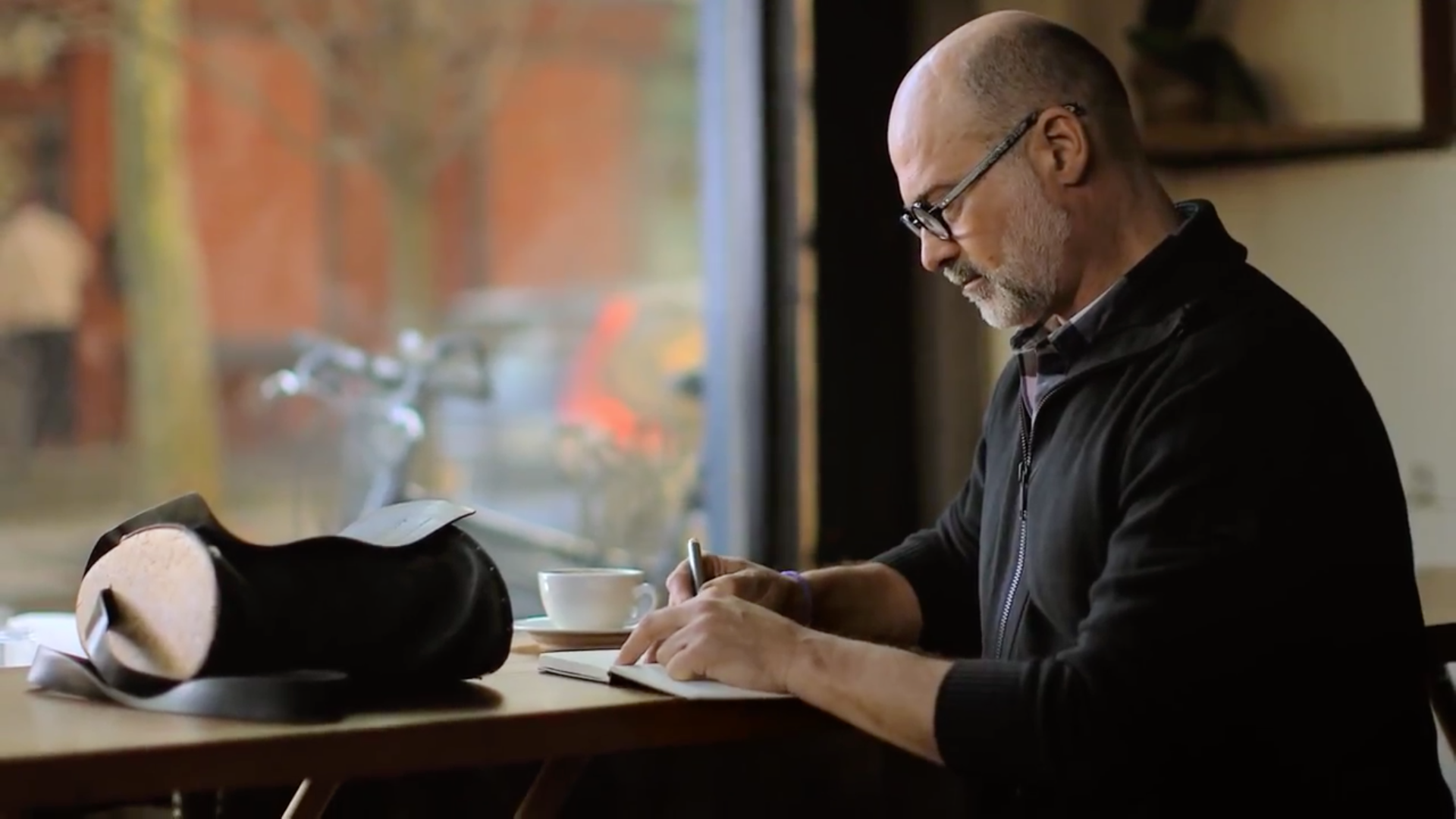 Bald man with glasses writing in a notebook at a cafe table, hat beside him, window view.