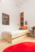 A minimalist bedroom with a wooden single bed, orange accents, wall posters, built-in bookshelf, and a red circular rug.