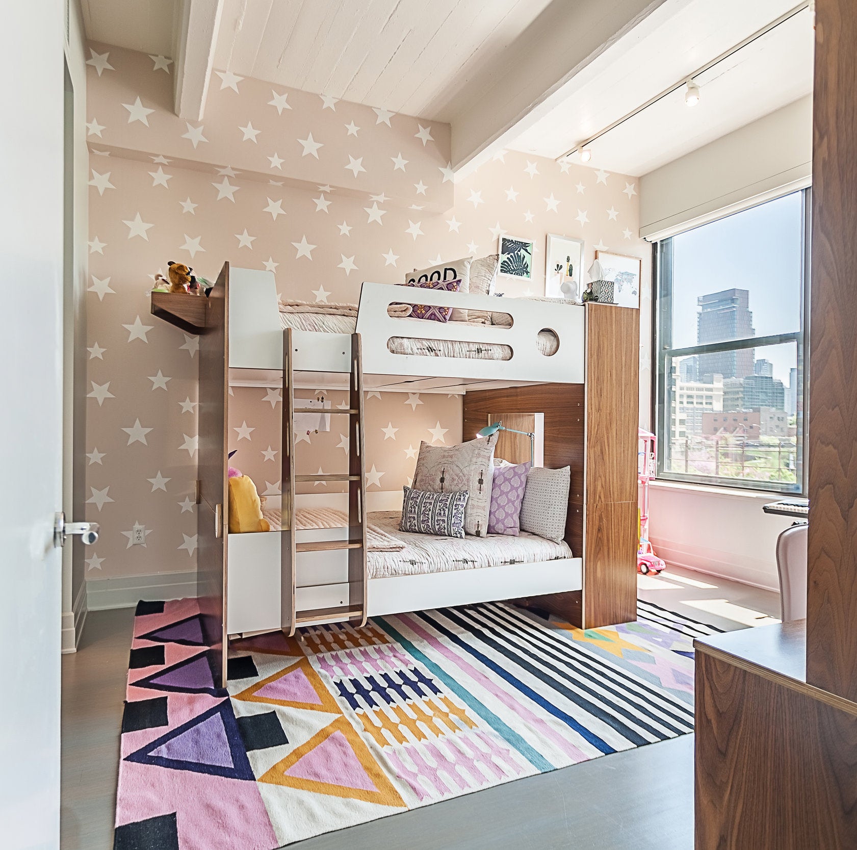 Bright children's room with bunk beds, star-patterned wallpaper, colorful rug, and city view window.