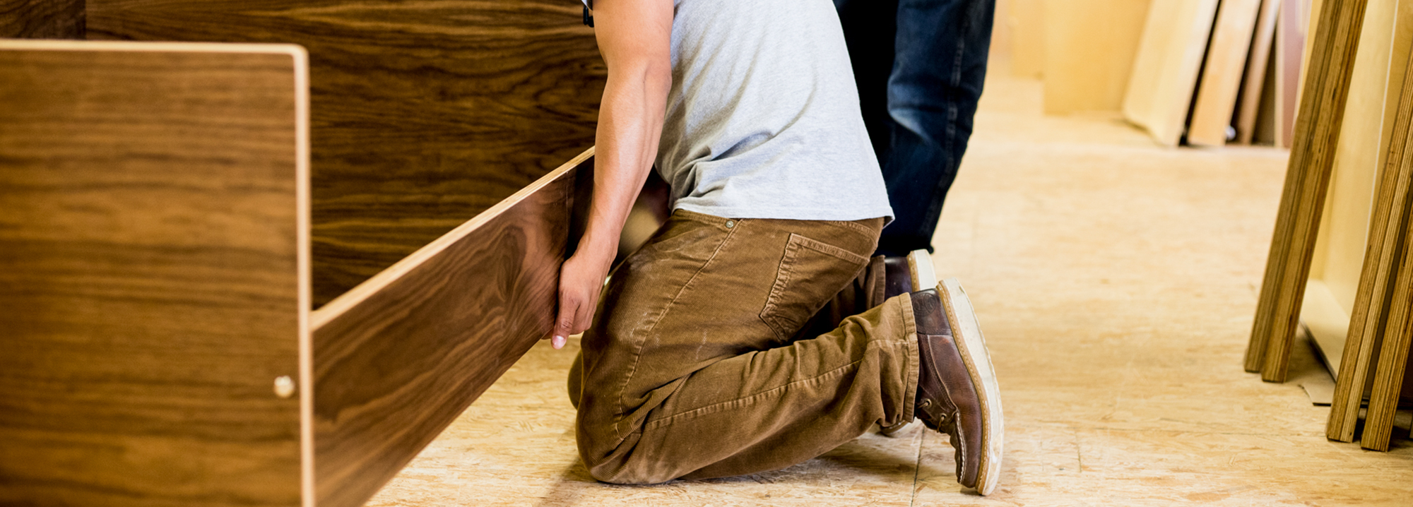 Person kneeling on the floor while assembling a wooden furniture piece in a workshop setting.