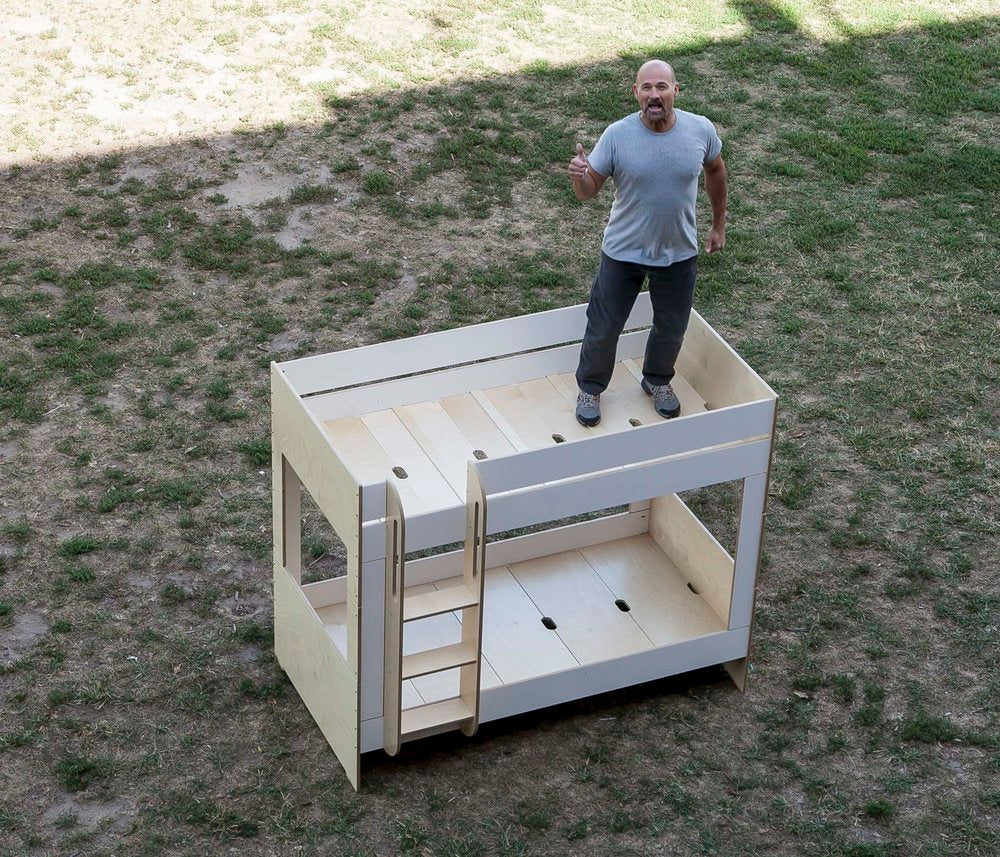 Aerial view of a man standing next to a small, wooden toddler bed with stairs on grass.
