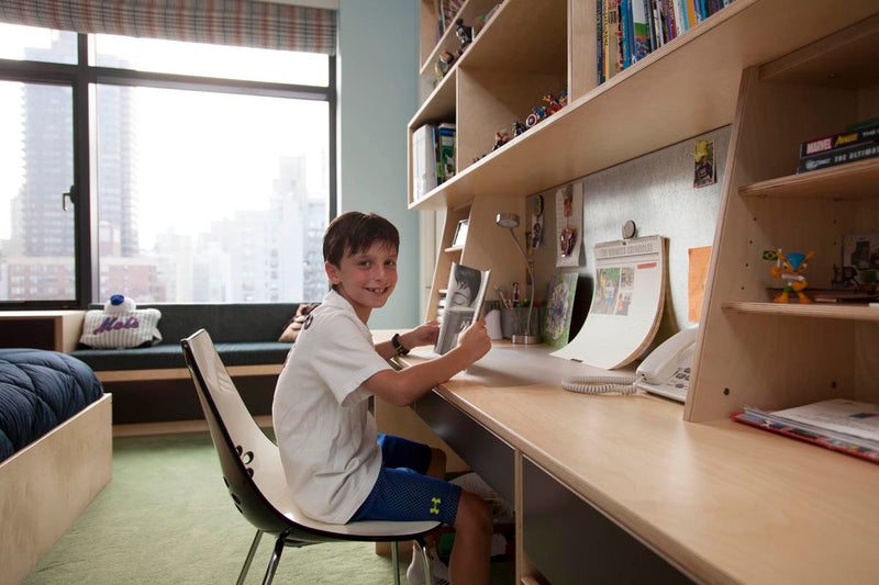 Young boy smiling and using a computer at a desk in a room with a city view and built-in shelves.