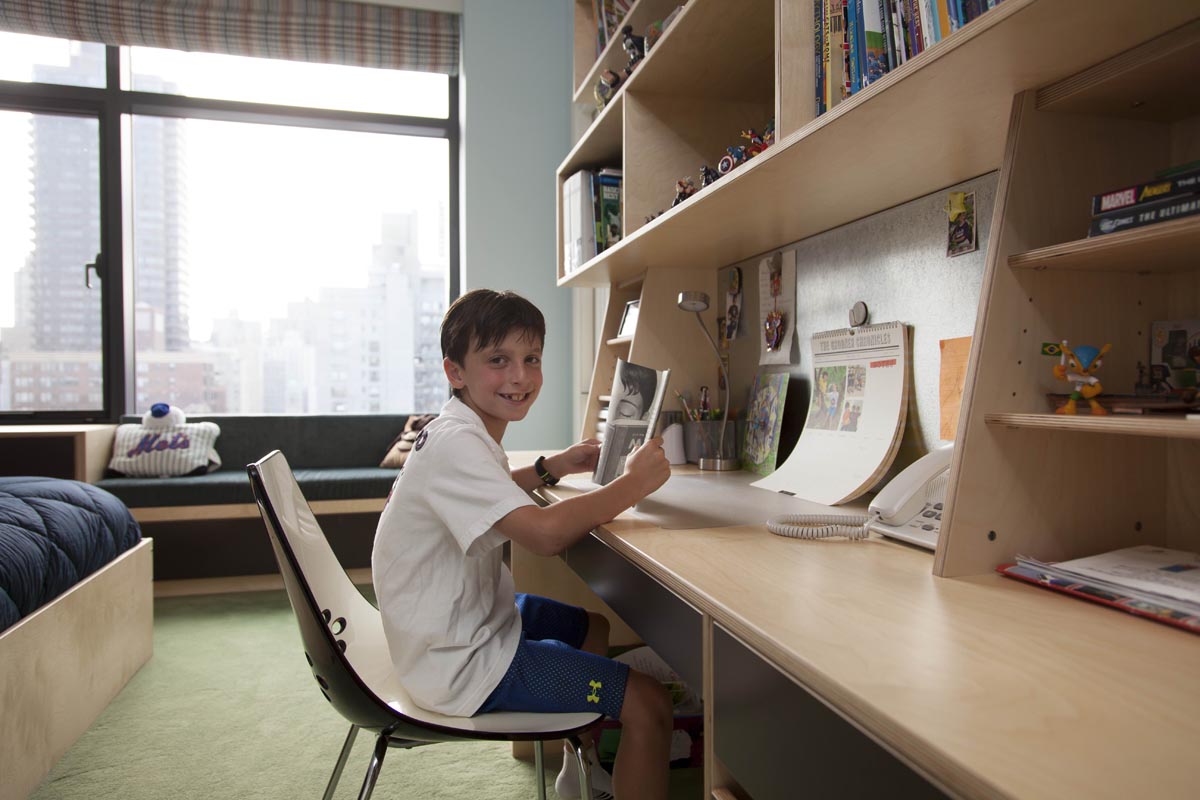 Young boy smiling and using a computer at a desk in a room with a city view and built-in shelves.