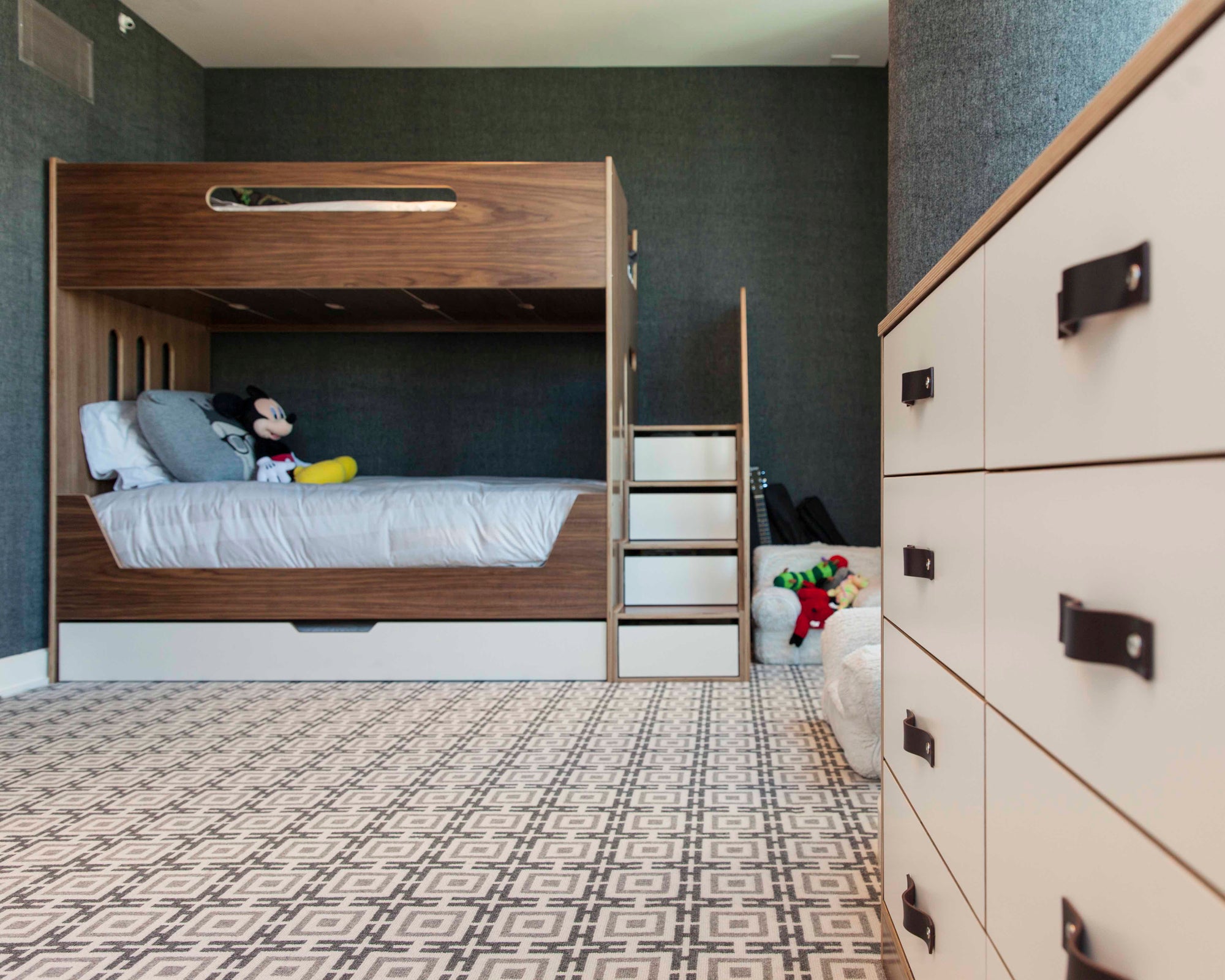 Children’s bedroom with bunk bed, storage cabinet, ladder, toys, and patterned carpet.