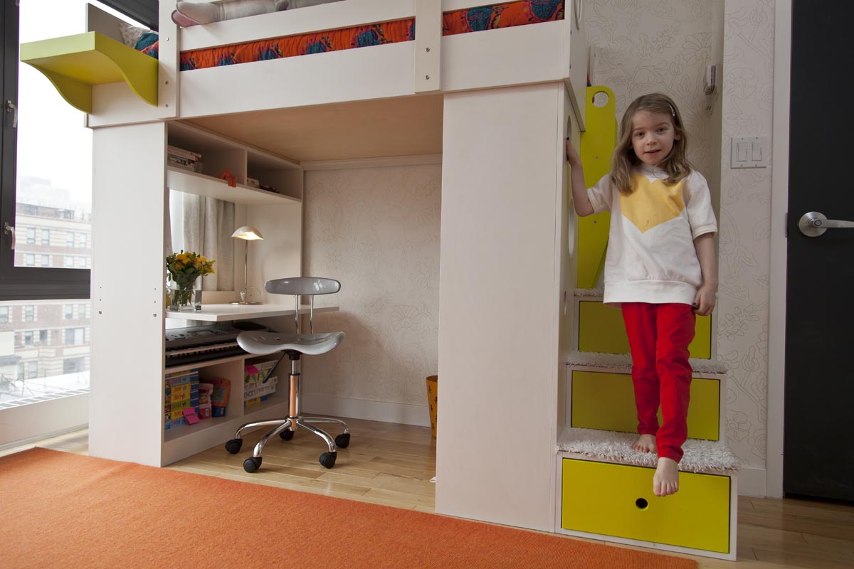 Girl in yellow top and red pants standing on yellow stairs of a loft bed, with a study area below.