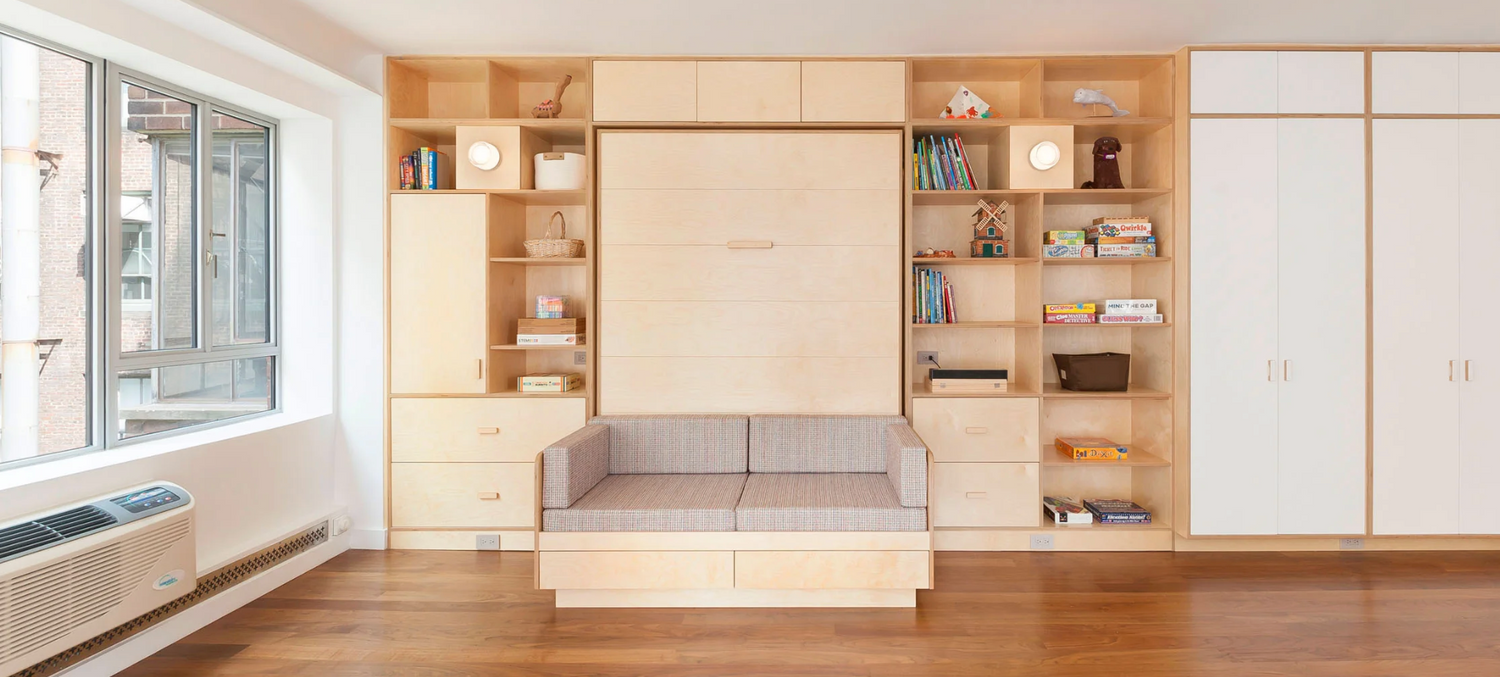 Bitch queen size Murphy tuck bed with grey couch cushions at base and custom matcign shelf and wardrobes built into the wall surrounding headboard in a white walled, wood floor room with a window on the left side.