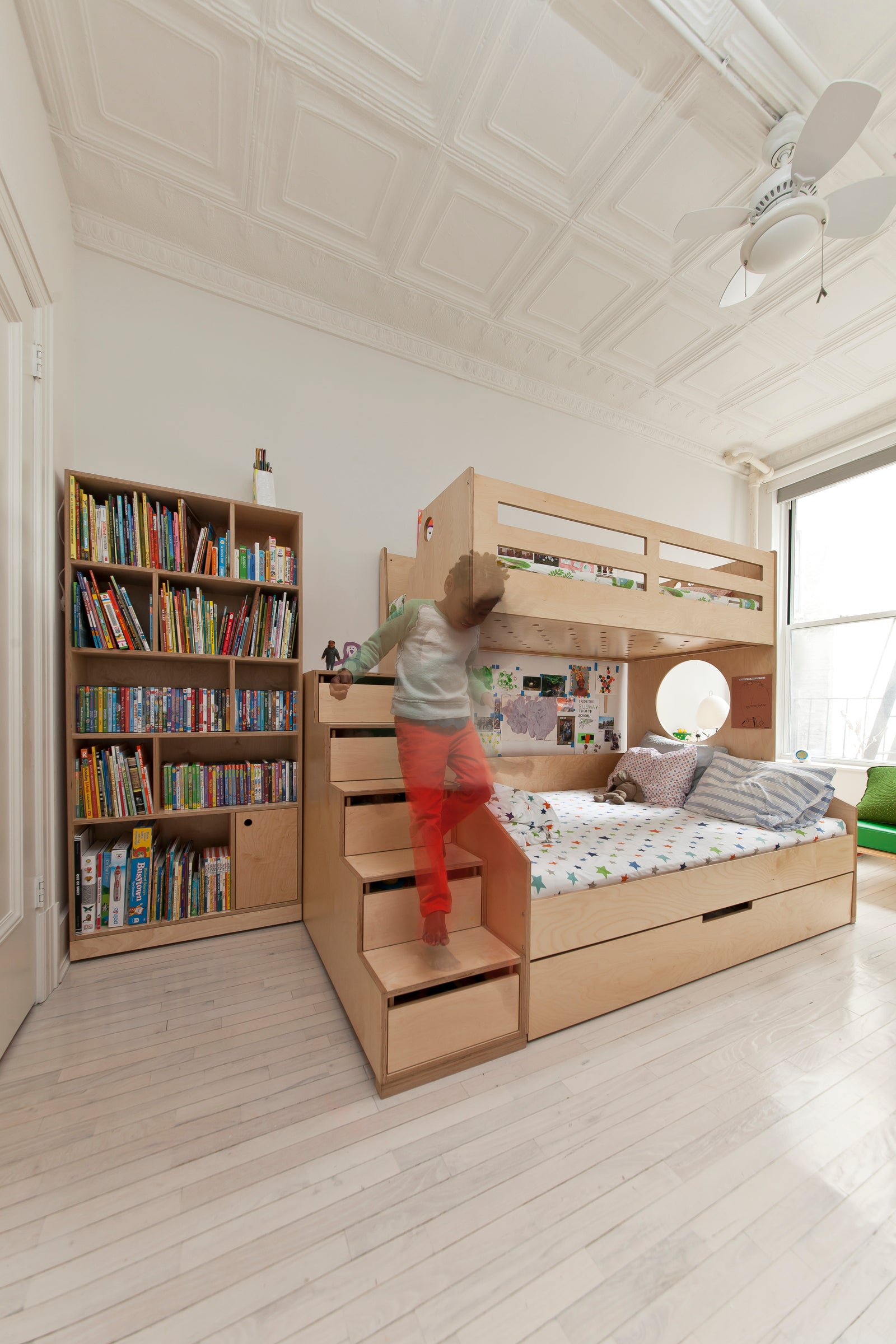 A child climbing steps to a wooden bunk bed in a room with white walls, hardwood floors, a bookshelf filled with books, and a ceiling fan.