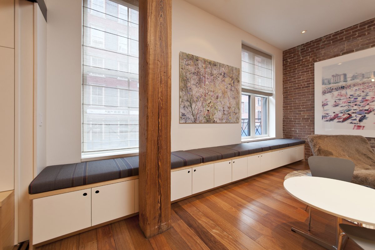 Urban living space with a long window bench, storage cabinets, exposed brick wall, and wooden beams.