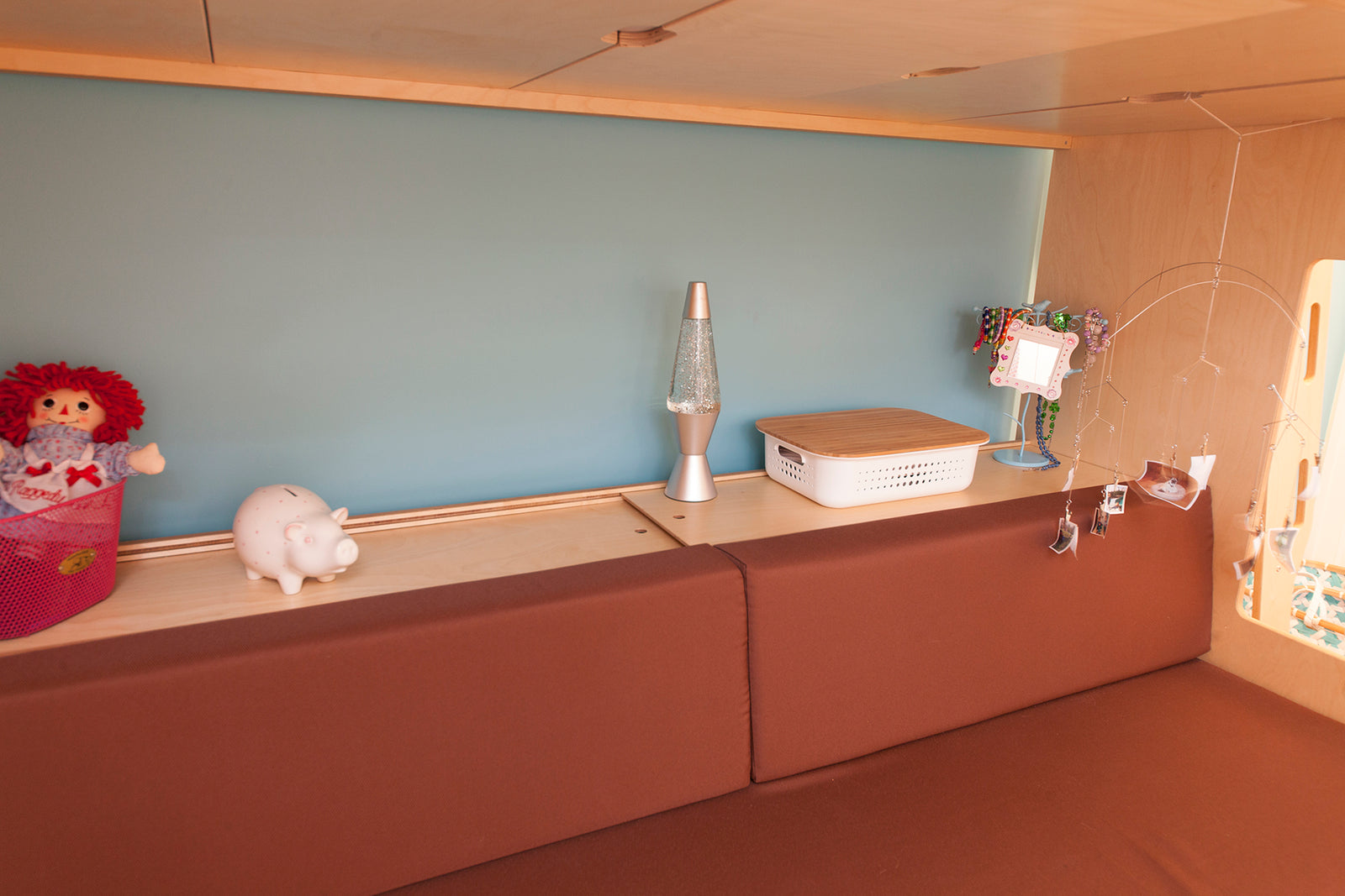 Shelf with a red rag doll, a piggy bank, a lava lamp, a white basket, and a lamp, with brown padded seating below.