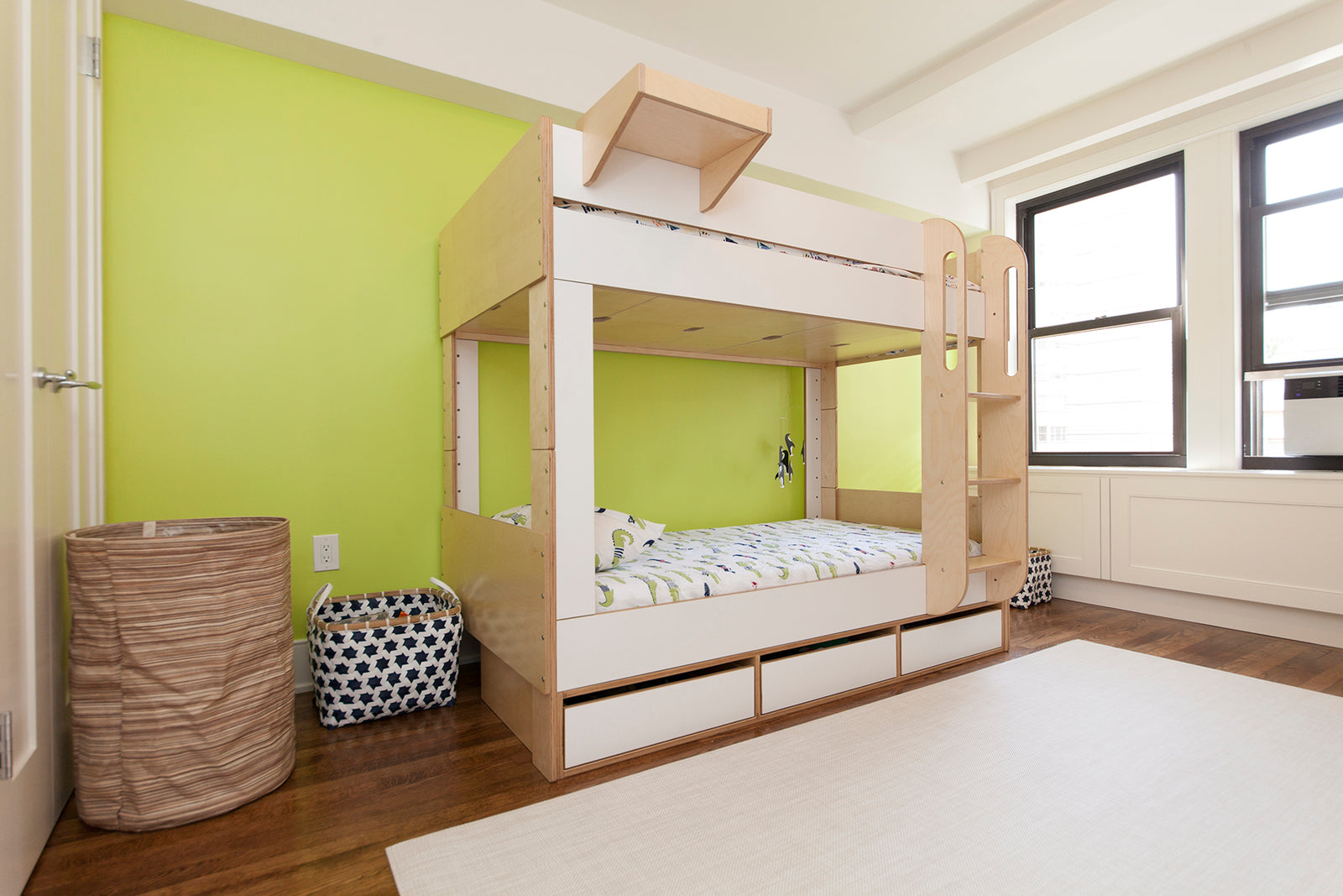 A wooden bunk bed with a ladder, storage drawers, and green accent wall, placed in a bright room with large windows.