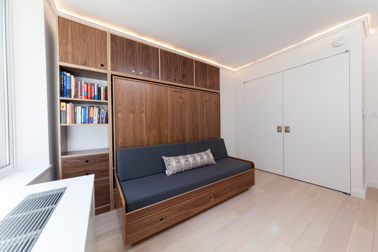 Modern room with wooden built-in cabinets, a daybed with cushions, LED lighting, and a bookshelf.
