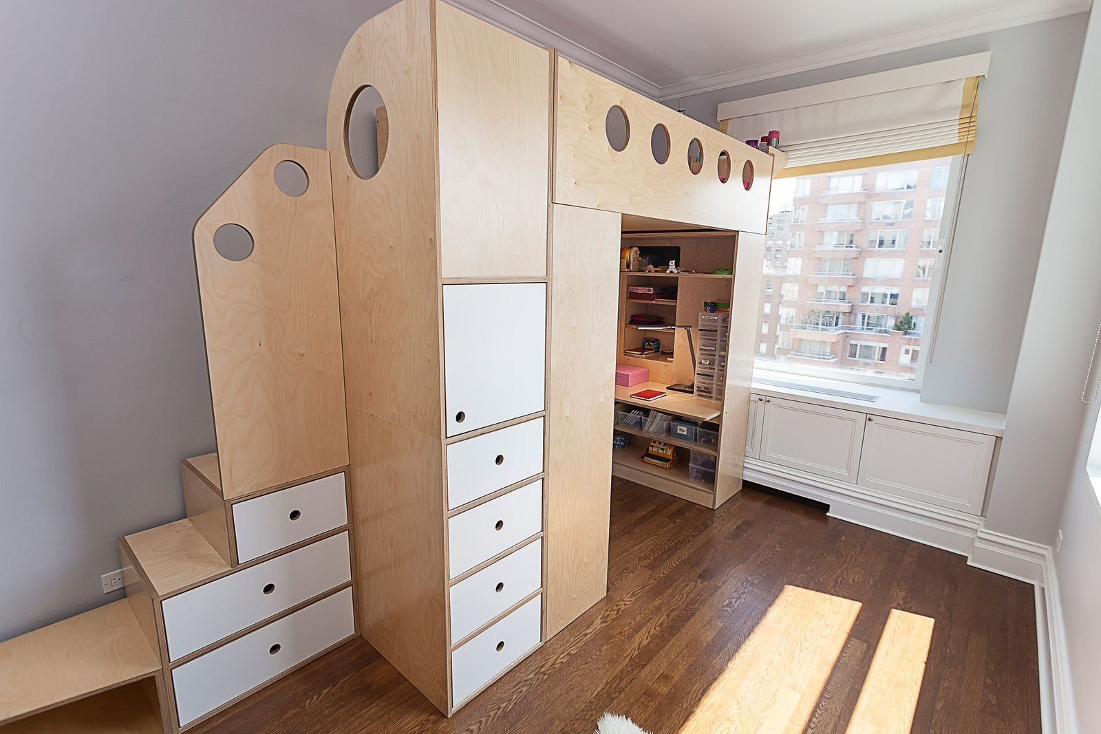 A wooden loft bed with storage drawers, a staircase, and a built-in desk beneath it, placed in a sunny room with large windows.