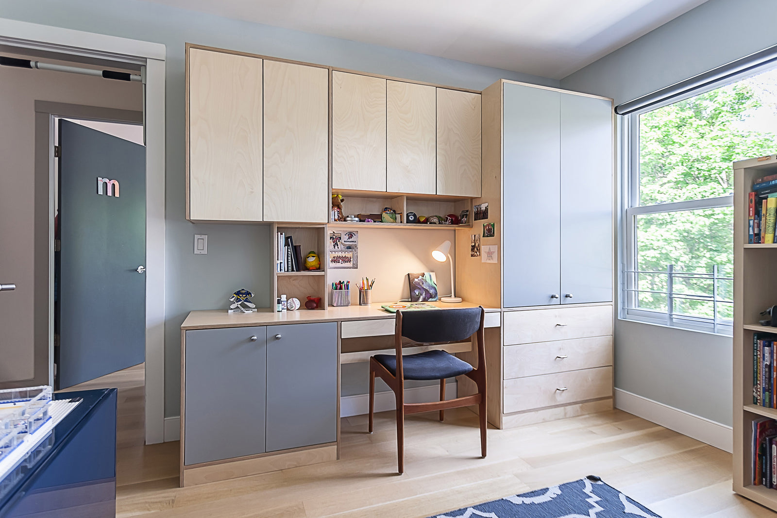 A modern kids' study room with built-in cabinets, a desk with a wooden chair, and a window providing natural light.