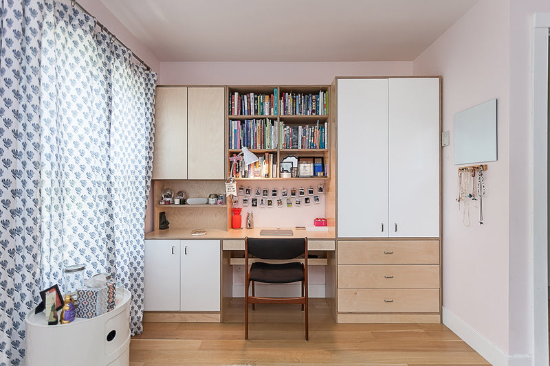 A children's study area with a wooden desk, bookshelves, and cabinets, a chair, and patterned curtains letting in natural light.