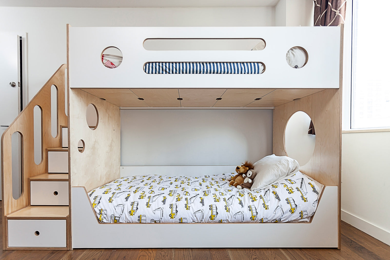 A modern wooden bunk bed with stairs and storage drawers, featuring circular cutouts, white bedding, and plush toys.