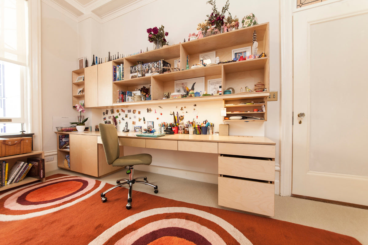 A spacious and organized study area with a long wooden desk, shelves filled with various items, and a green chair on a rug.