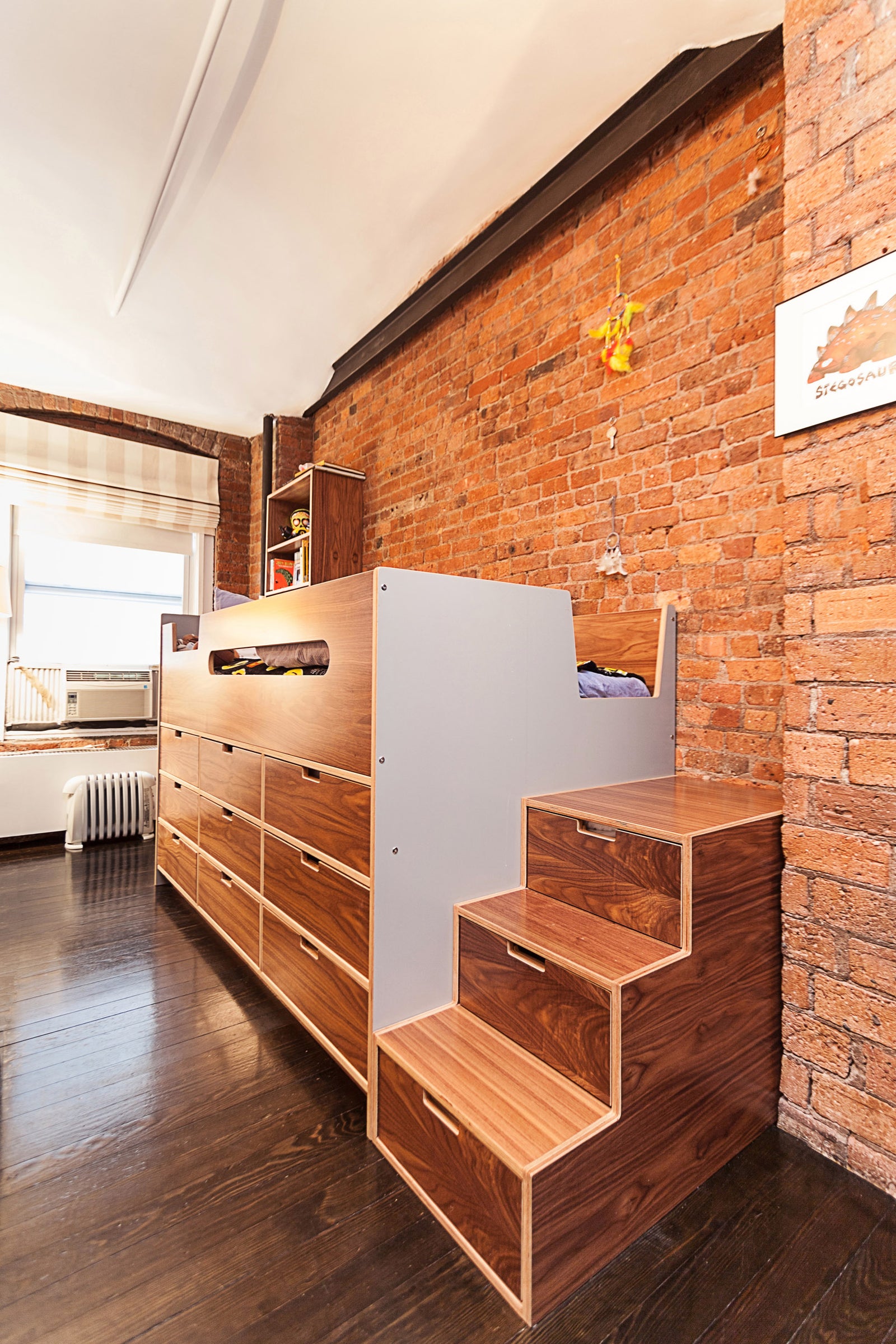 A stylish kids' room with a bunk bed featuring wooden drawers, a brick accent wall, and stairs with built-in storage drawers.