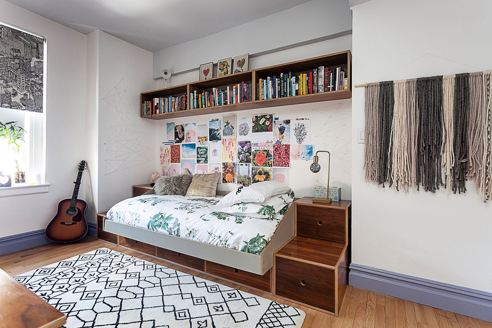 Bedroom with a daybed, floral bedding, bookshelf above, art-covered wall, guitar in the corner, and geometric rug on the floor.