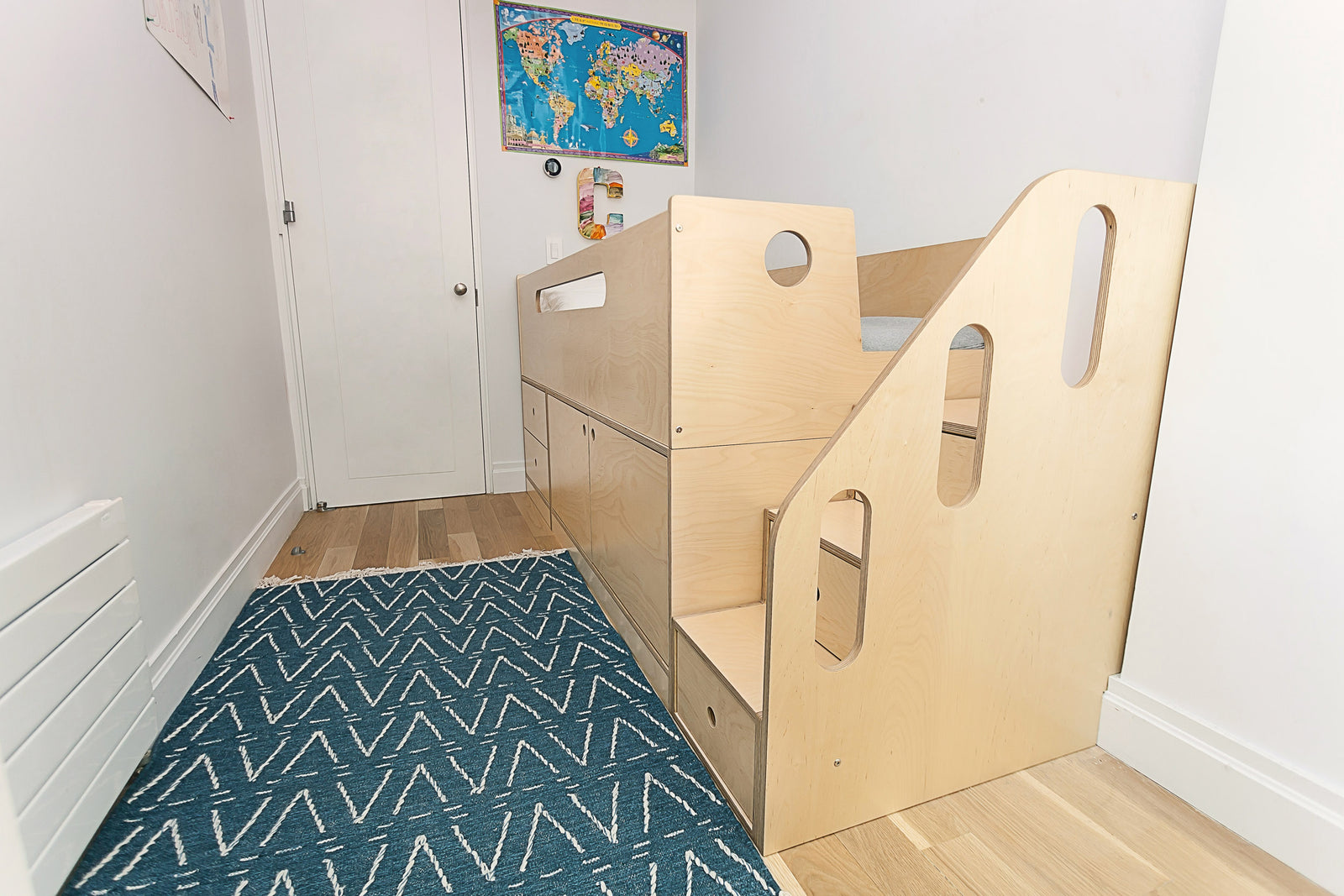  A wooden loft bed with stairs and storage in a narrow room, featuring a blue rug and a world map on the wall.