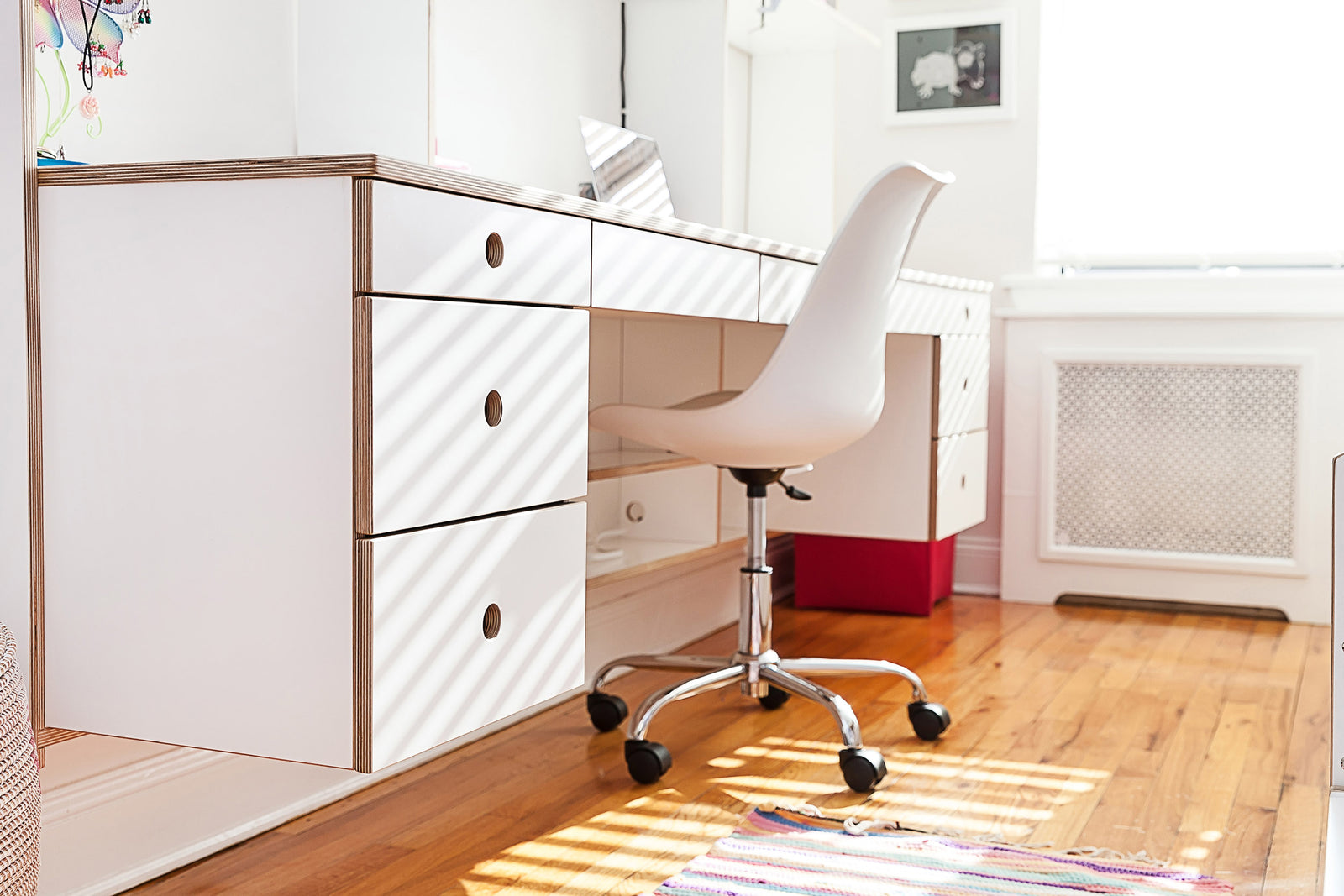 A modern study area with a white desk, drawers, a swivel chair, and sunlight streaming through the window onto a wooden floor.
