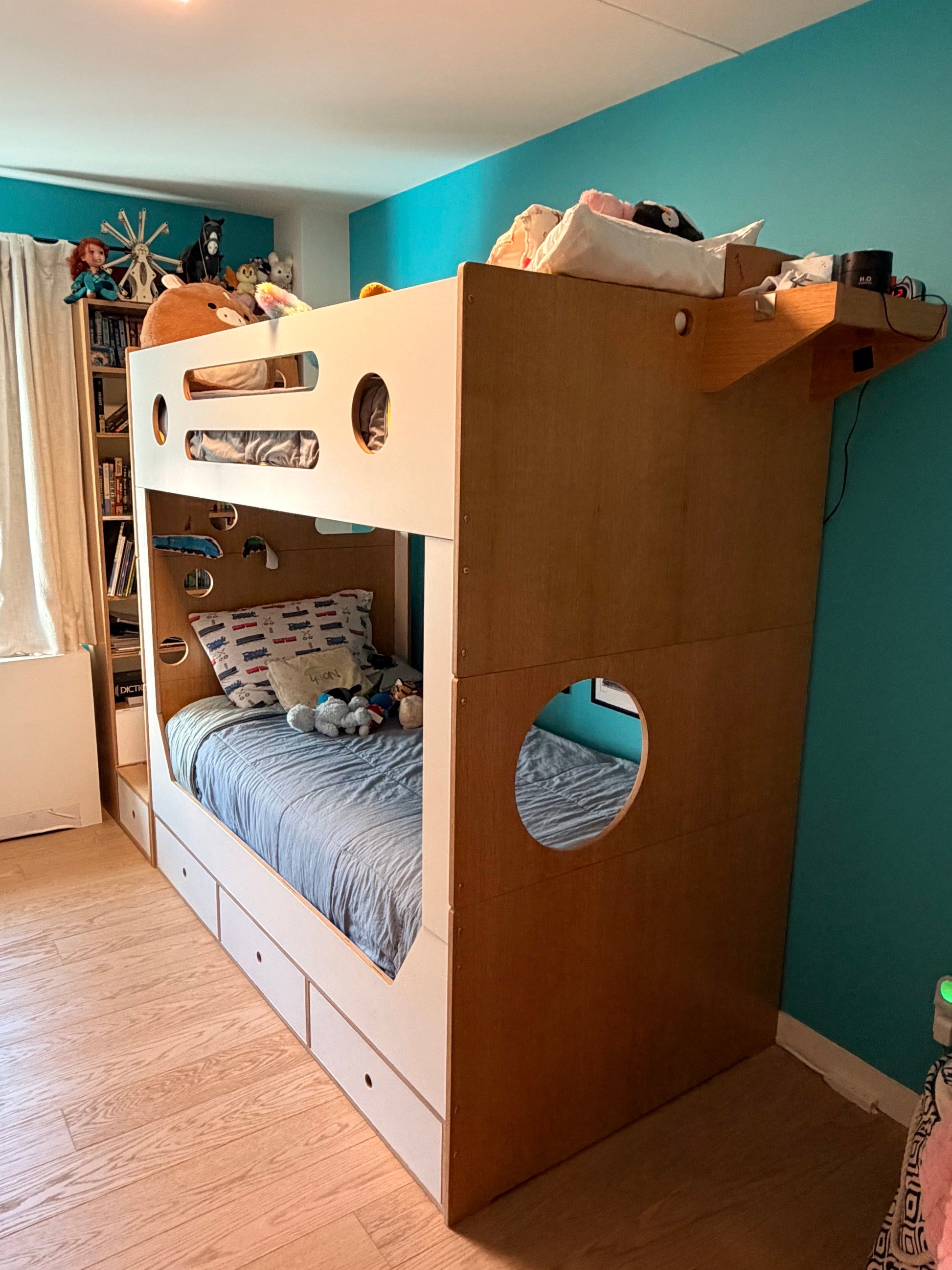 A large wooden bunk bed with a white top bunk frame and blue bedding, shown in a child's bedroom with teal walls and light wood flooring.