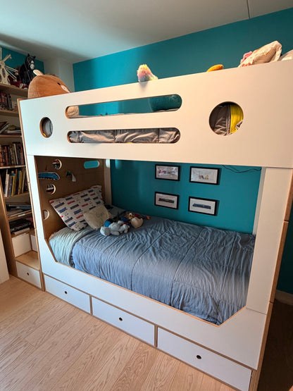 A white wooden bunk bed with a lower storage drawer bed, shown in a child's bedroom with a teal wall and light wood flooring.