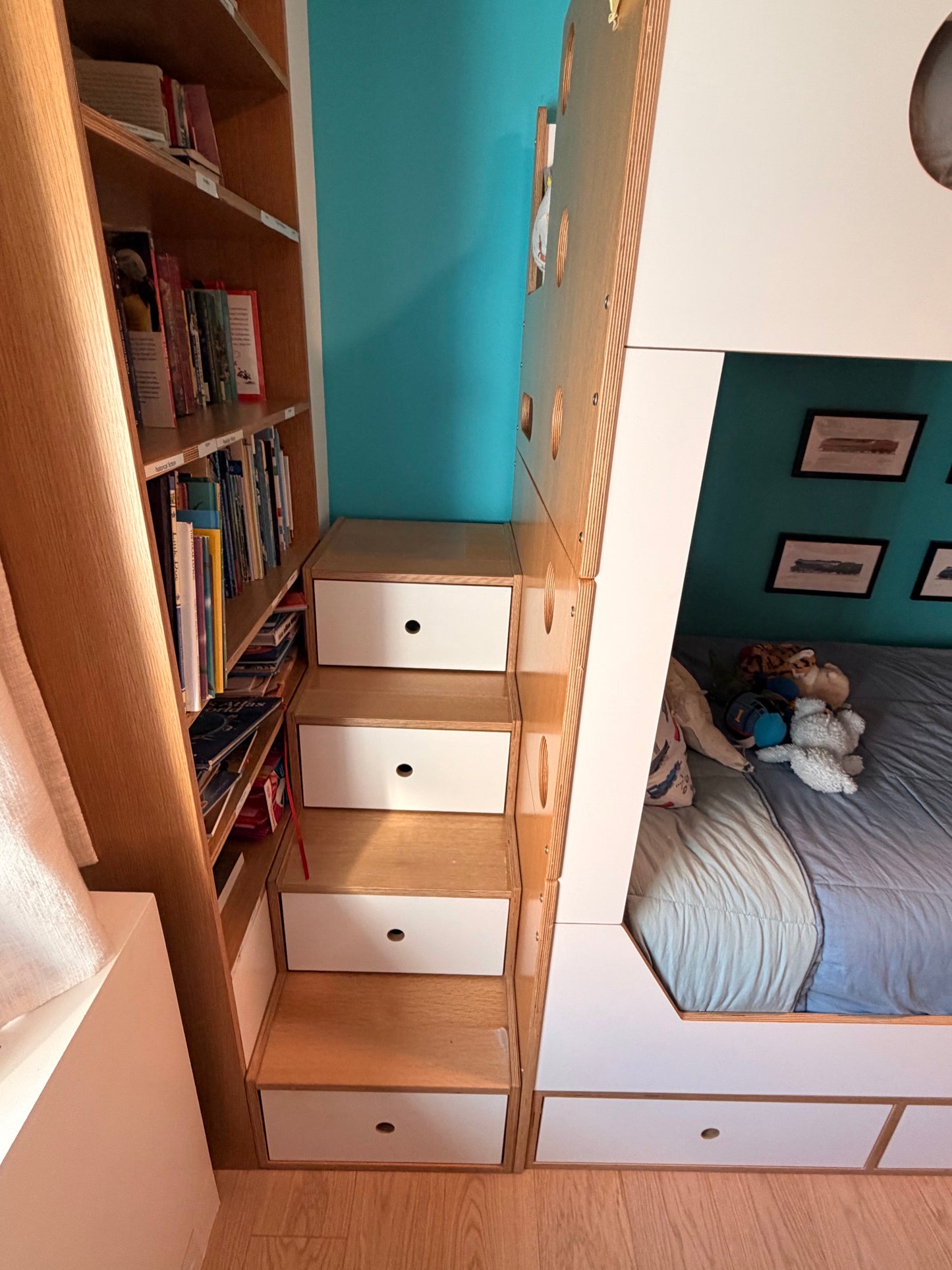 A compact wooden bunk bed with integrated storage drawers and a built-in bookshelf on the left side, shown in a bedroom setting next to a teal wall and bed.