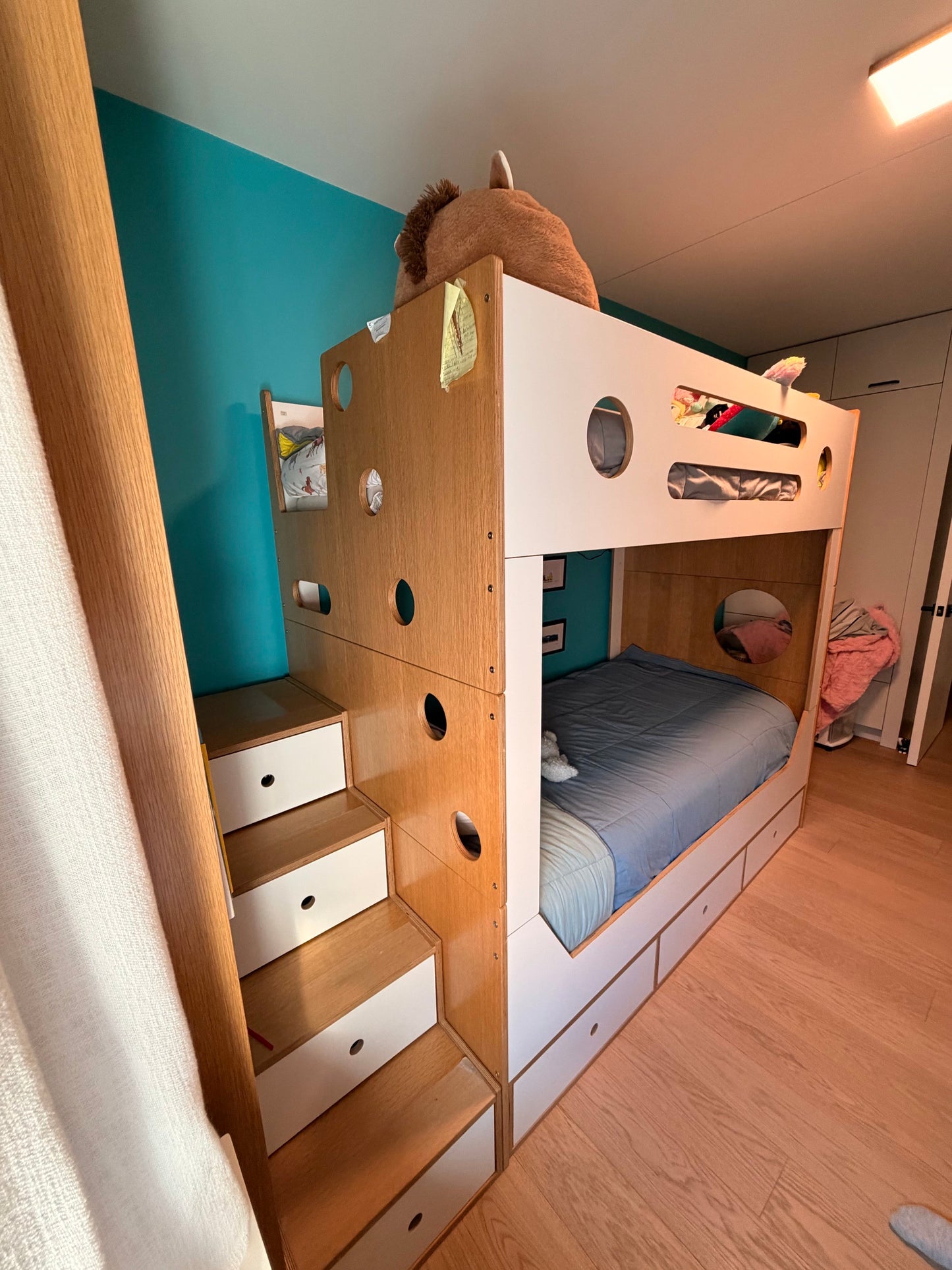 A compact wooden and white bunk bed with integrated stairs and storage drawers, shown in a child's bedroom with a teal wall and light wood flooring.