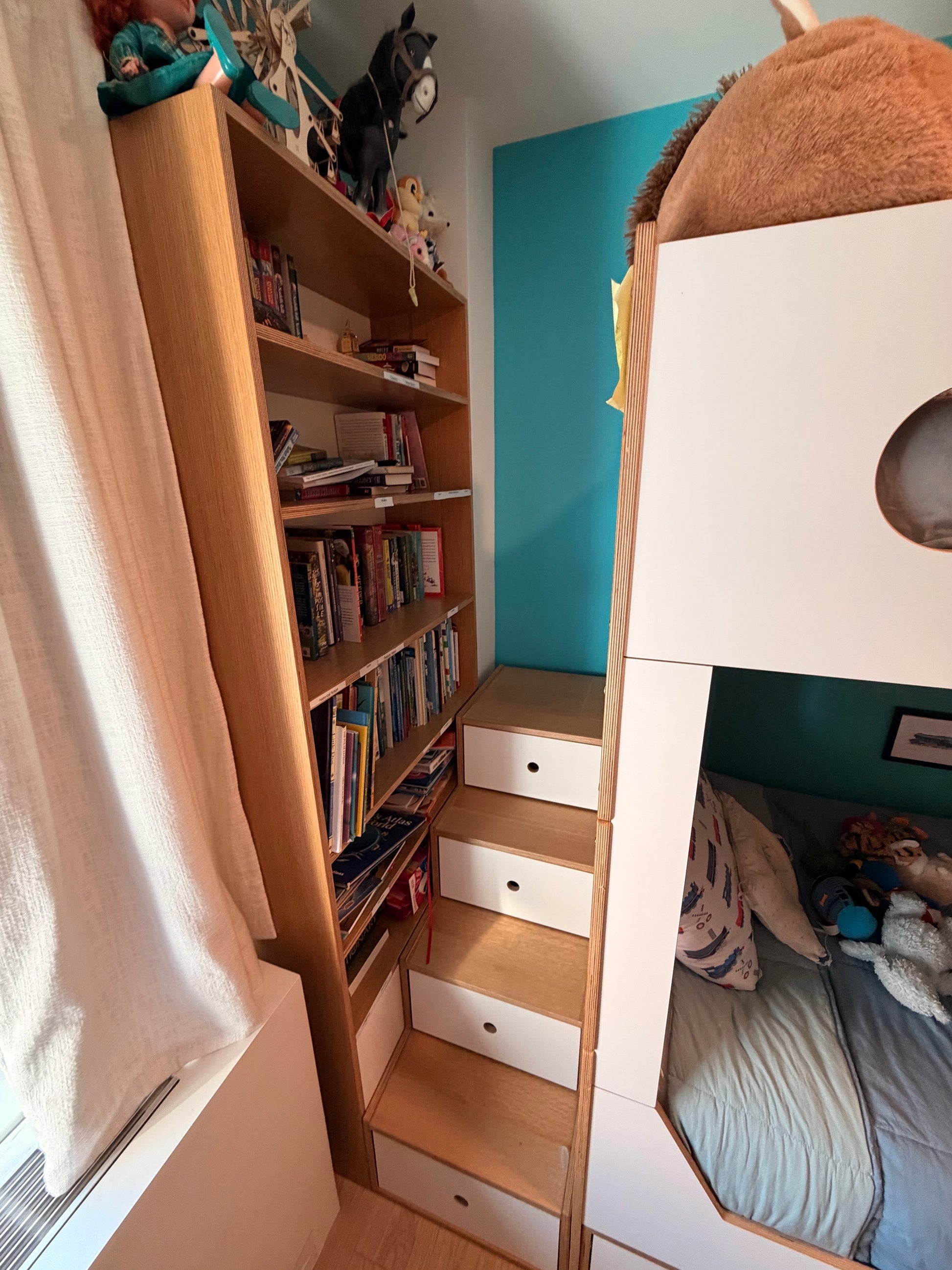 A narrow bedroom corner with a tall light wood bookshelf filled with books, built-in steps with white drawers, and part of a white bunk bed with a blue wall behind it.