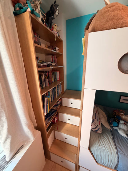 A narrow bedroom corner with a tall light wood bookshelf filled with books, built-in steps with white drawers, and part of a white bunk bed with a blue wall behind it.