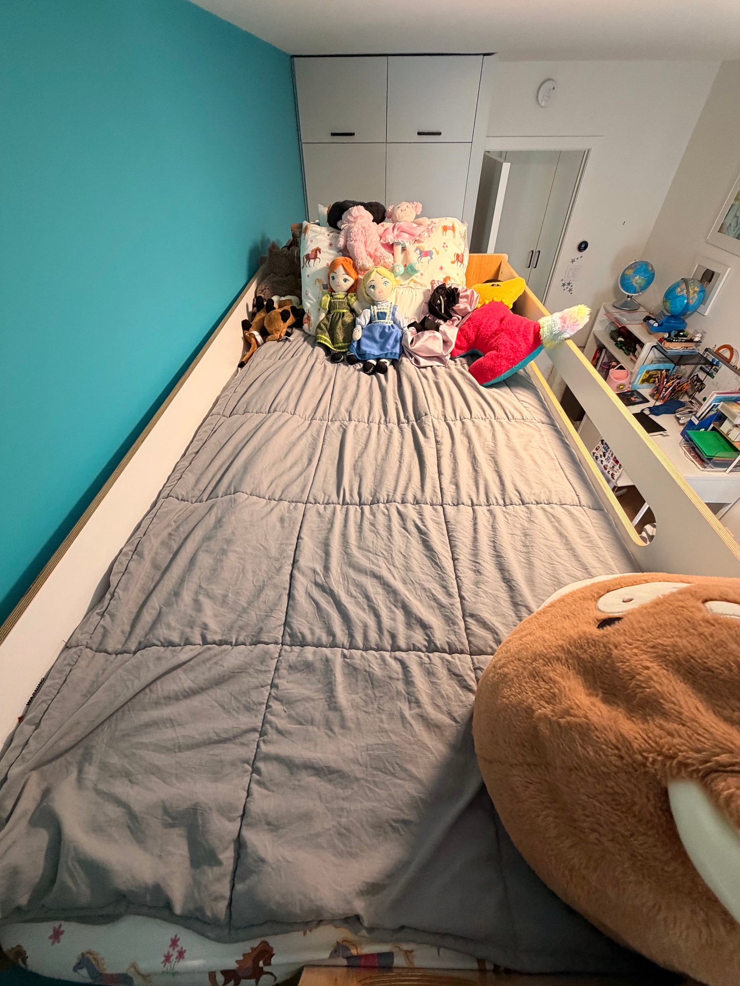 A high-angle view of a bed in a child's bedroom, featuring a gray quilted comforter, a colorful patterned pillow, and various plush toys. The room has a prominent teal blue wall on the left, white walls and wardrobe in the background, and a large brown plush toy in the foreground.