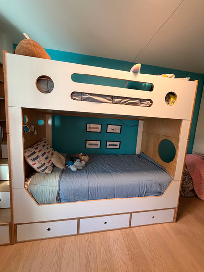 A white and light wood bunk bed with a modern, playful design, featuring circular and horizontal cutout details on the top rail and built-in storage drawers underneath. The bed is styled with blue bedding and toys, set against a teal accent wall in a child's bedroom.