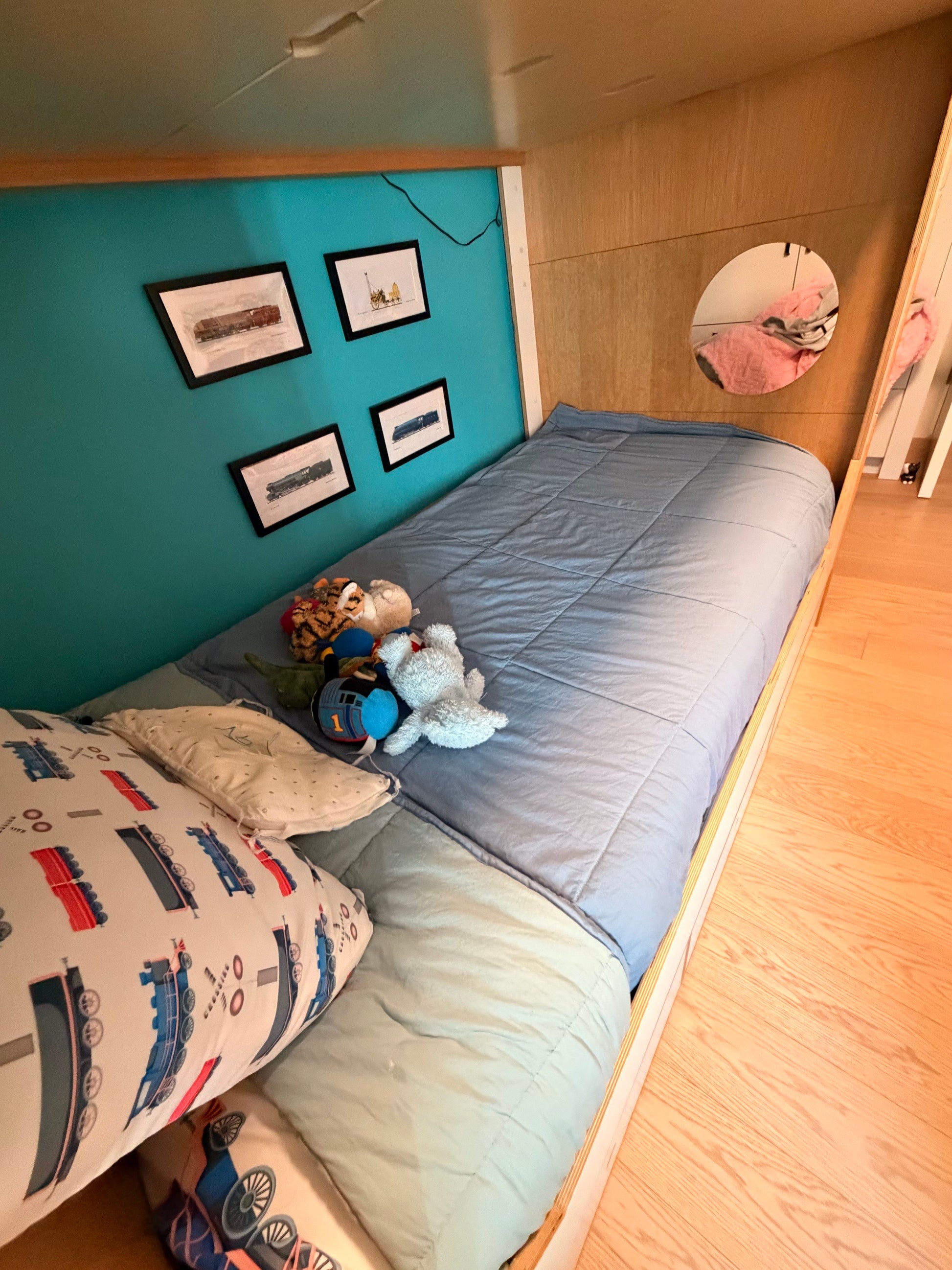 A children's single bed with a light blue quilted duvet and a light green sheet, decorated with a blue and white train-patterned pillow, plush toys, and framed pictures on a teal wall.
