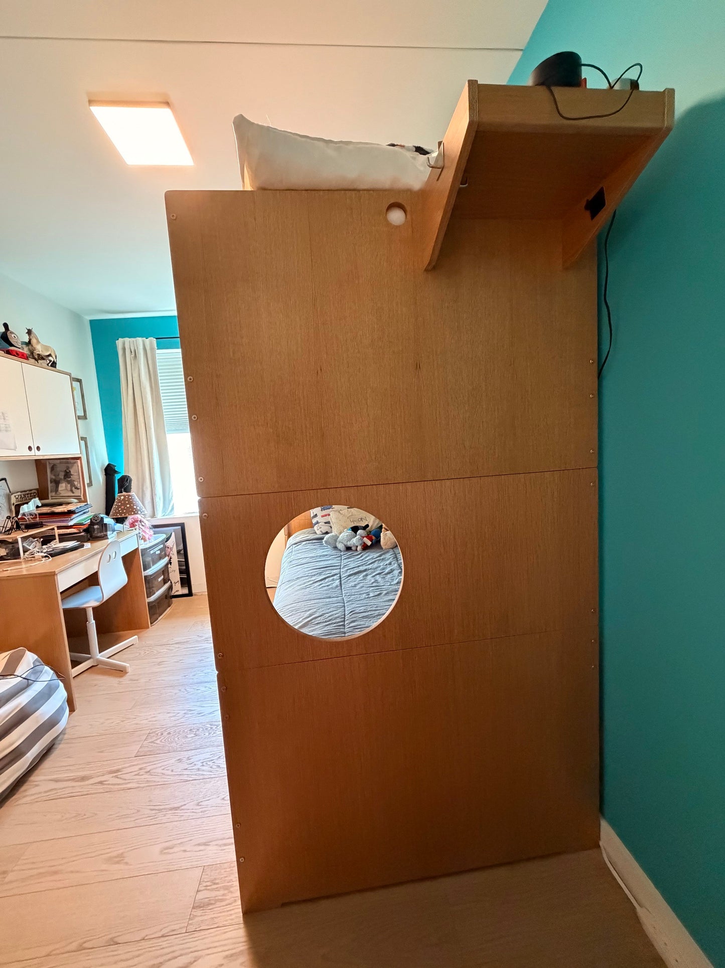 A tall wooden bed frame or storage unit with a built-in shelf on top and a circular cutout in the front, shown in a bedroom with a teal wall and light wood floor.