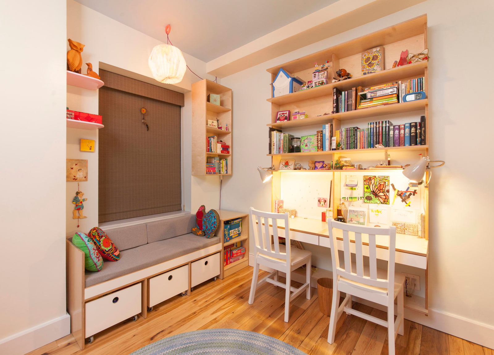 A cozy study area with dual desks, white chairs, shelves with books and toys, and a seating nook with storage drawers below.