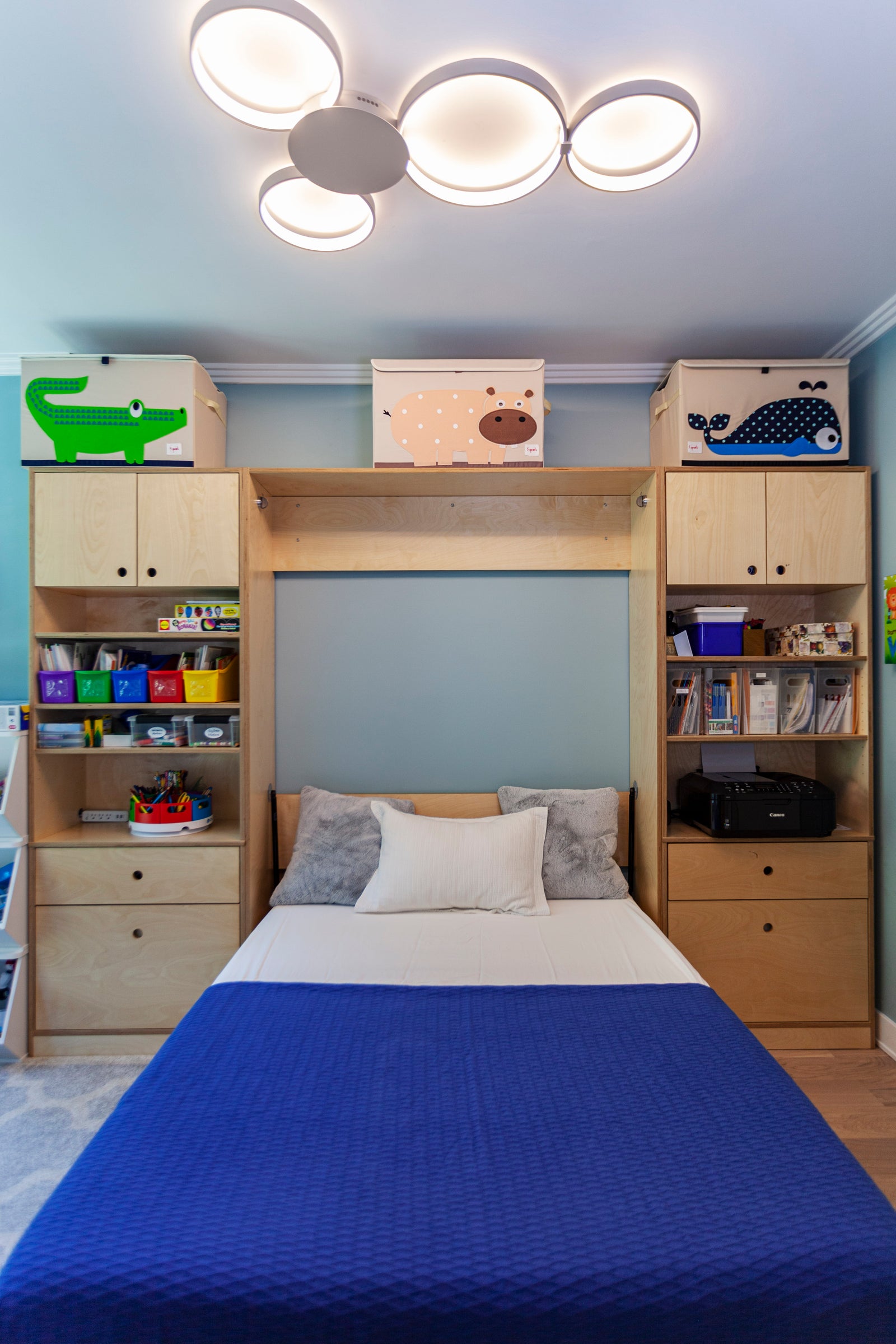 A tidy bedroom with a double bed, blue bedspread, built-in shelving, toy storage boxes, and a modern ceiling light fixture.