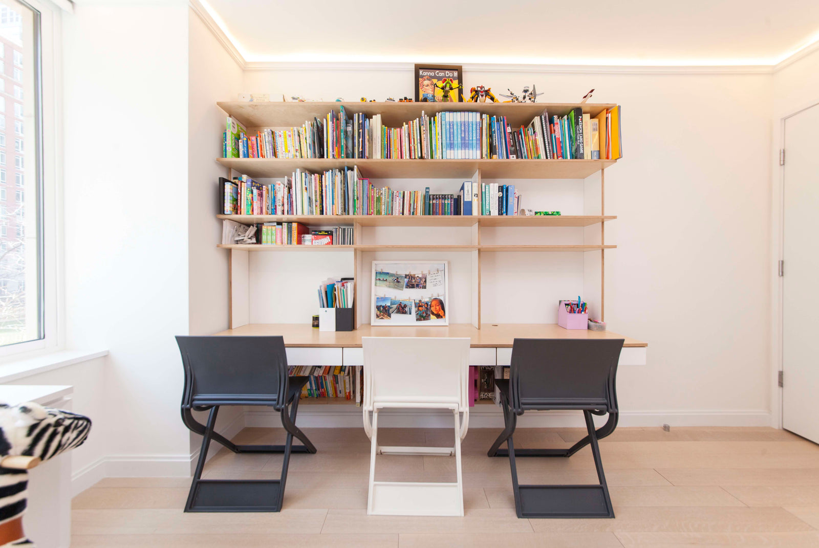 A bright study area with a triple desk, three chairs, and shelves filled with books and toys, illuminated by ceiling lights.